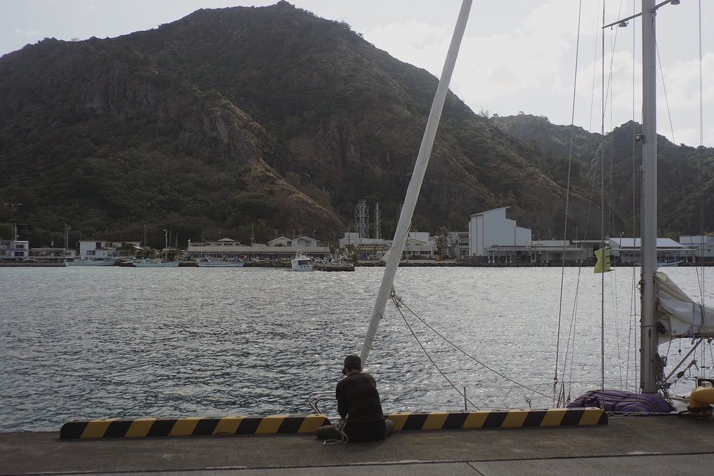A person sitting on the concrete wall of a fishing harbor, tying the dock lines of a sailboat. The tide is low so only the mast is visible, flying a yellow quarantine flag
